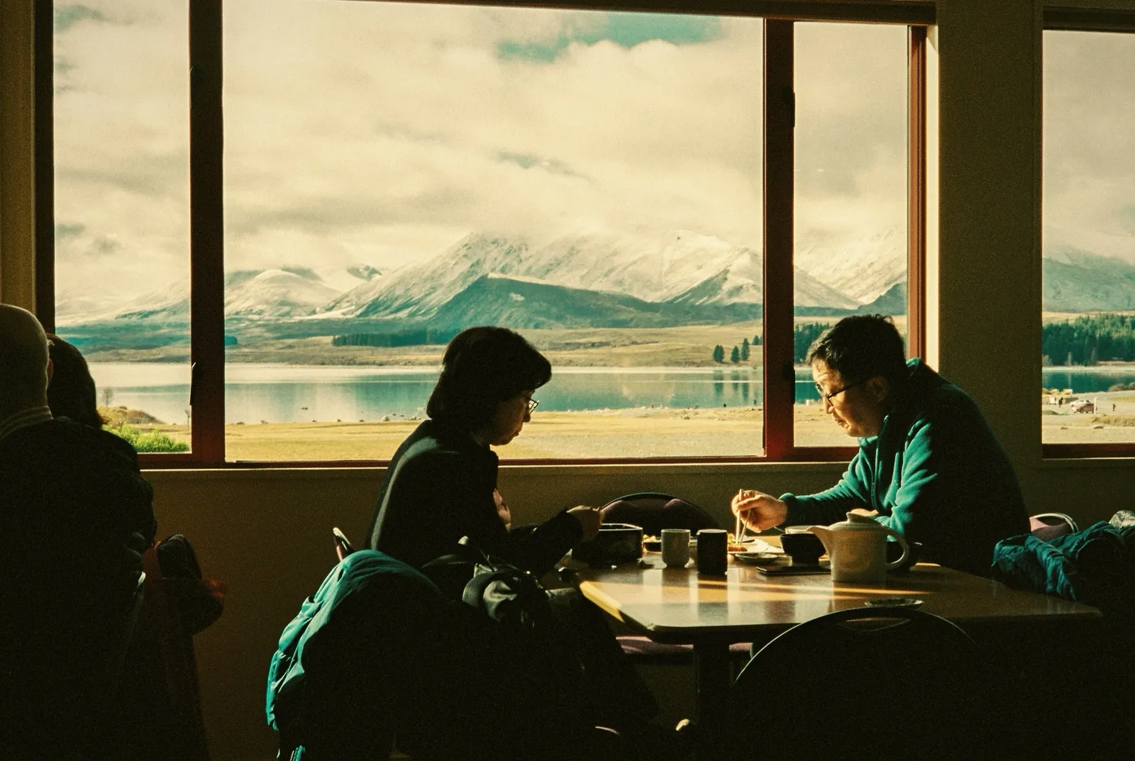 Two people at a cafe table with a lake and snow-capped mountains through the window